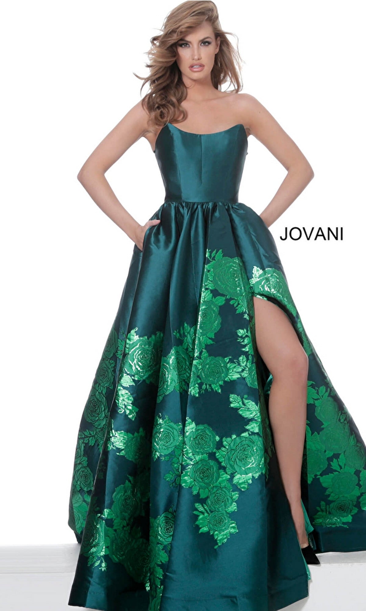 A woman stands against a plain white background wearing the Jovani Strapless Floral Long Prom Ball Gown 02038, featuring shiny green floral patterns and a high slit. The word